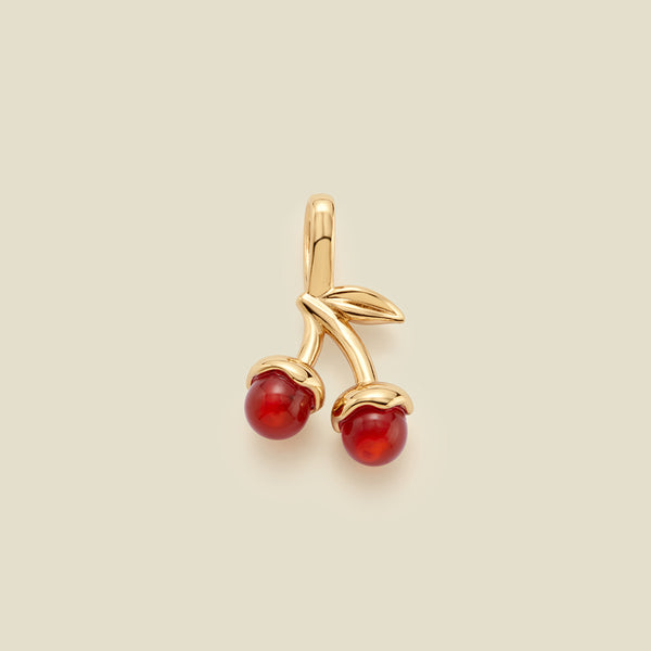 A cherry-shaped jewelry charm in polished gold-tone metal with two red agate stone cherries and small gold leaves, centered on a light beige background.