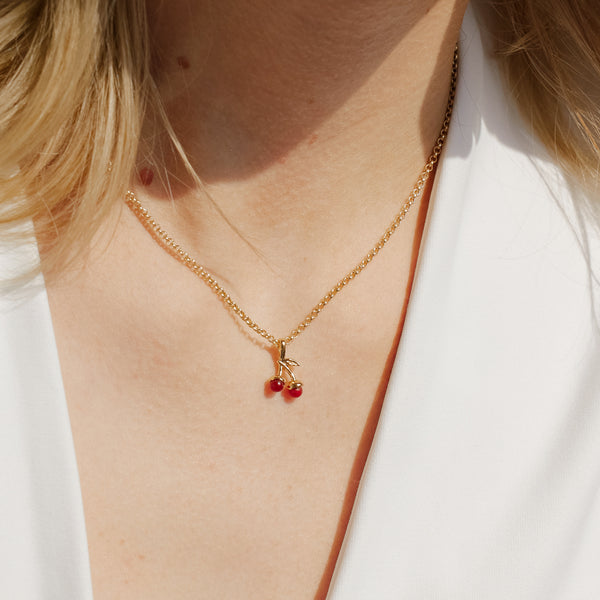 A close-up of a person in a white top wearing a delicate gold chain necklace with a cherry charm featuring two red agate cherries and gold leaves.