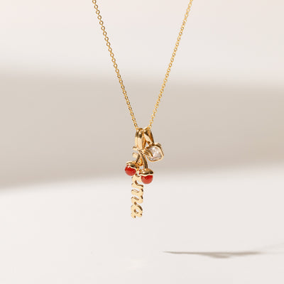 A delicate gold chain necklace holding a vertical script 'mama' charm, a cherry charm with two red agate stones, and a square clear gemstone charm, shown dangling in front of a solid white background with natural shadows.