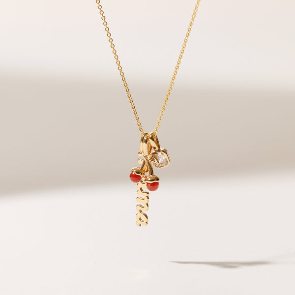 A delicate gold chain necklace holding a vertical script 'mama' charm, a cherry charm with two red agate stones, and a square clear gemstone charm, shown dangling in front of a solid white background with natural shadows.