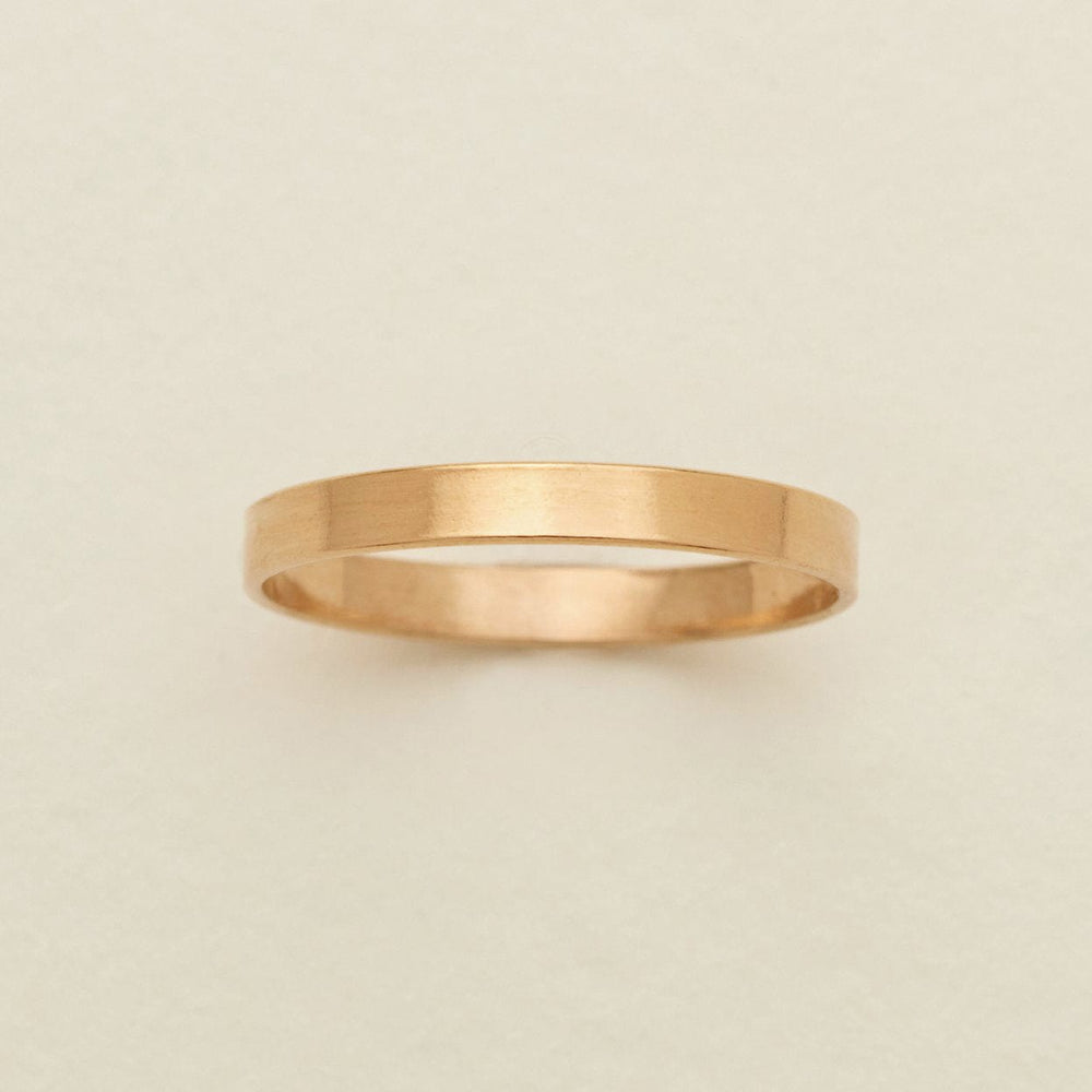 A minimalist, thin gold band ring with a brushed texture, centered on a light beige background.