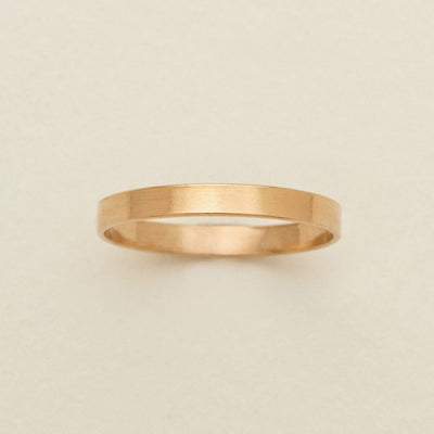 A minimalist, thin gold band ring with a brushed texture, centered on a light beige background.