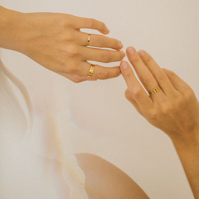 A person's hands wearing multiple of the same simple gold band ring stacked on different fingers, shown on a light beige background.