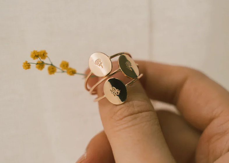 A hand holding three dainty gold rings with large oval discs, each with a different floral design engraved, held up against a white back drop with a yellow flower.