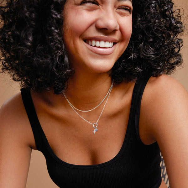 A woman in a black tank top in front of a solid light brown background, wearing a flat wide silver necklace layered with a silver round link chain necklace with a silver ring in the center. On the ring is a silver cross charm and a square light blue gemstone charm.