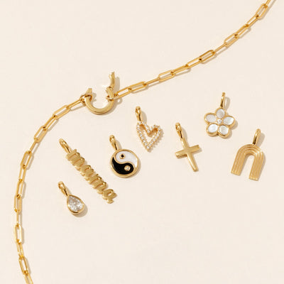 A gold paperclip link chain necklace with a round ring in the center, showing an open latch to add charms. Around the necklace are multiple gold charms, including a clear gemstone pear cut charm, a 'mama' vertical charm in a script font, a yin yang charm, heart charm, cross charm, flower charm, and rainbow charm.