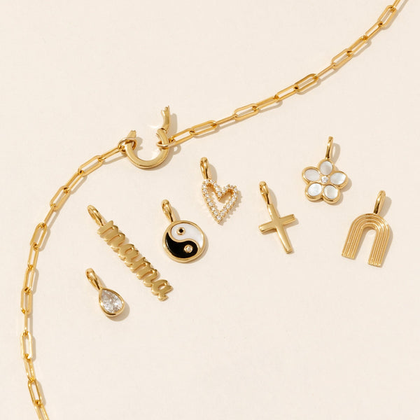 A gold paperclip link chain necklace with a round ring in the center, showing an open latch to add charms. Around the necklace are multiple gold charms, including a clear gemstone pear cut charm, a 'mama' vertical charm in a script font, a yin yang charm, heart charm, cross charm, flower charm, and rainbow charm.