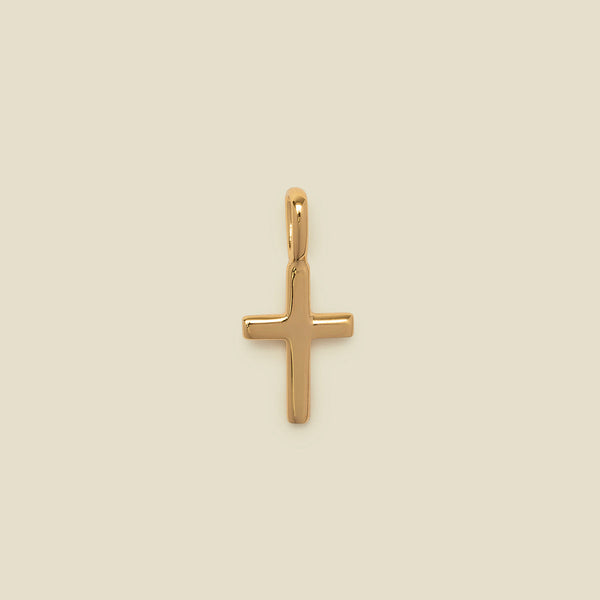 A polished gold cross pendant with a smooth, rounded finish and a top loop, centered on a solid, warm beige background.