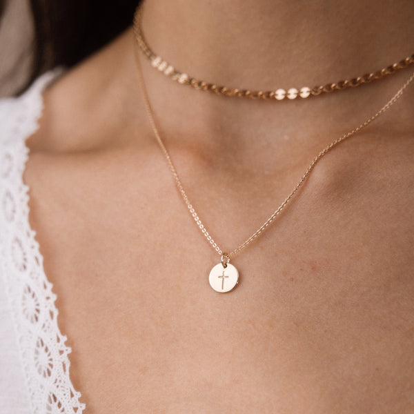 A close-up of a person's neck and upper chest wearing layered gold necklaces, including a delicate chain necklace with a small round disc pendant engraved with a cross.