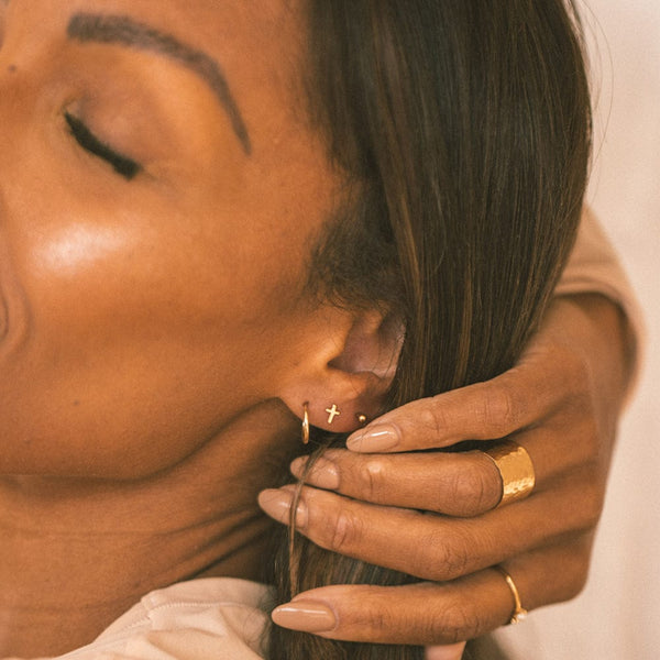 A woman's ear wearing a gold hoop earring, a gold cross stud and a gold round bead stud earring.