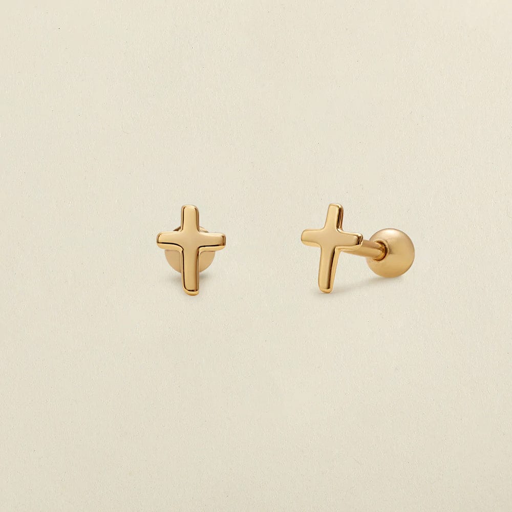 A pair of gold cross stud earrings shown on a solid light beige background, with one earring facing forward and the other angled to show the post and backing.