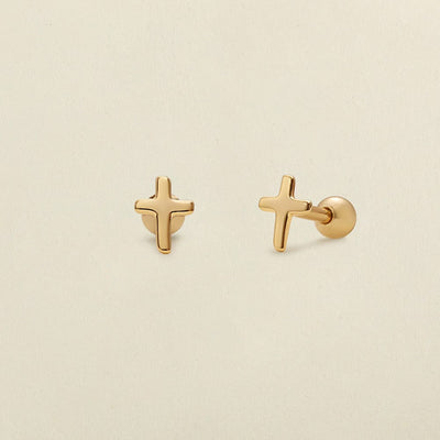 A pair of gold cross stud earrings shown on a solid light beige background, with one earring facing forward and the other angled to show the post and backing.