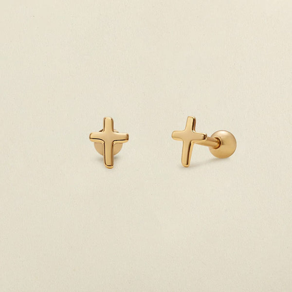 A pair of gold cross stud earrings shown on a solid light beige background, with one earring facing forward and the other angled to show the post and backing.
