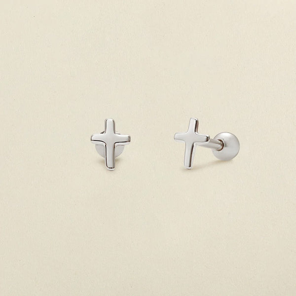 A pair of minimalistic silver cross stud earrings with round ball backings, shown on a solid light beige surface.