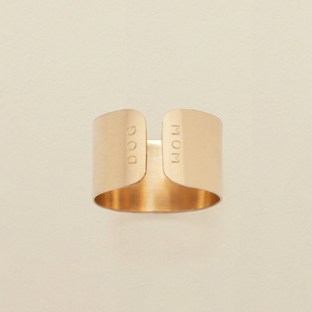 A gold cigar band ring with an engraving on both sides of the opening, reading 'DOG' and 'MOM', shown on a solid light beige background.