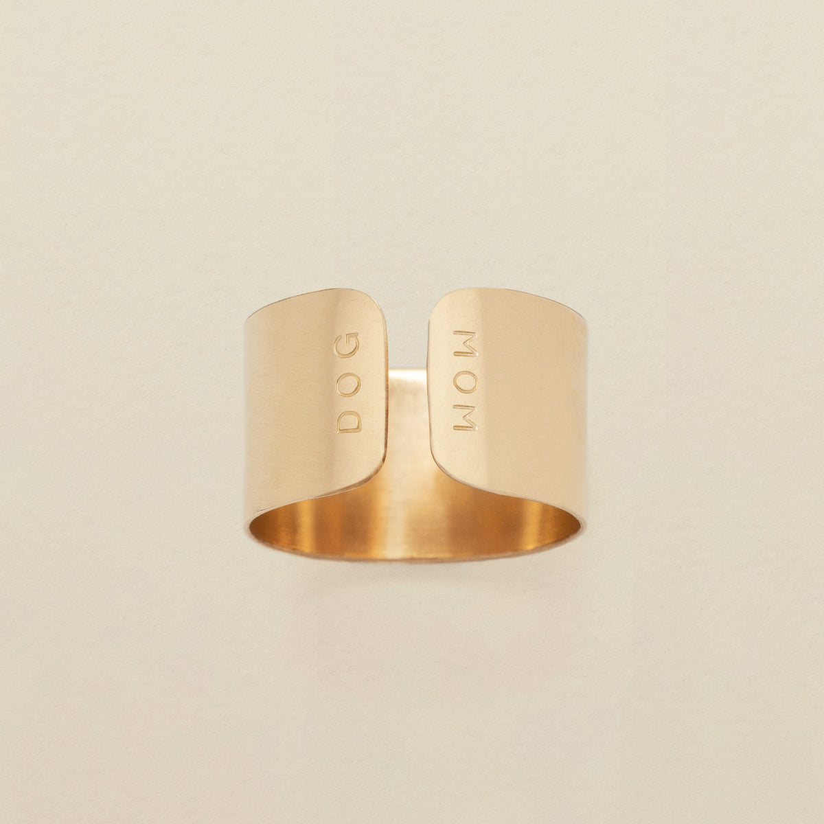 A gold cigar band ring with an engraving on both sides of the opening, reading 'DOG' and 'MOM', shown on a solid light beige background.