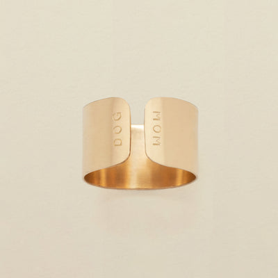 A gold cigar band ring with an engraving on both sides of the opening, reading 'DOG' and 'MOM', shown on a solid light beige background.