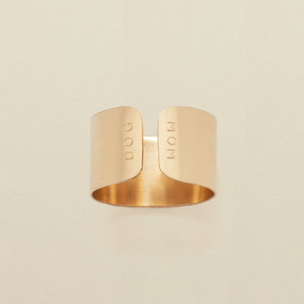 A gold cigar band ring with an engraving on both sides of the opening, reading 'DOG' and 'MOM', shown on a solid light beige background.