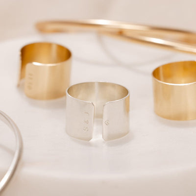 A silver polished cigar band ring with two gold cigar band rings blurred in the background, shown on a solid white surface. The silver ring is engraved on both sides of the opening, reading 'S & J' on one side and a heart on the other.