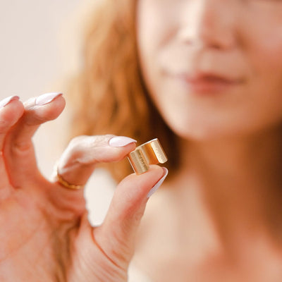A person's fingers holding a gold wide cigar band ring, showing the opening.