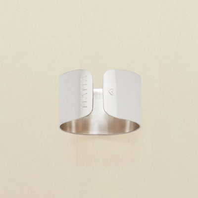 A polished silver cigar band ring with engraving on both sides of the opening, reading 'MAMA' on one and a heart on the other. The ring is shown on a solid light beige background.