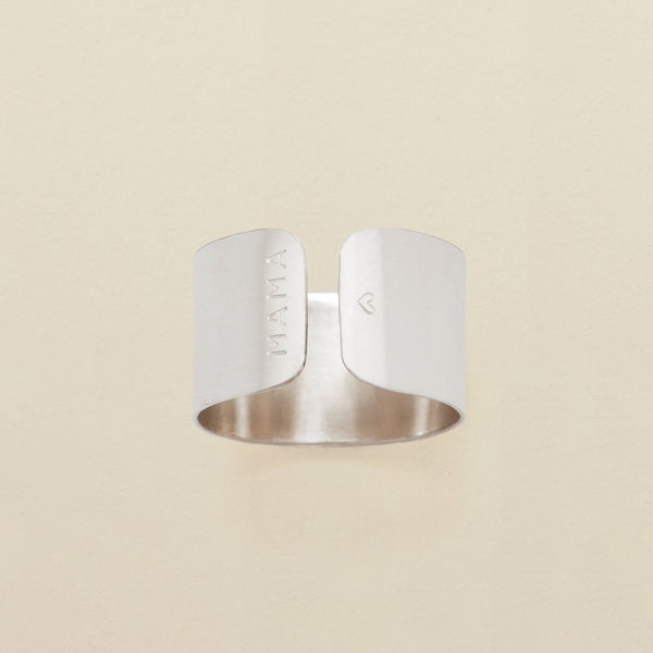 A polished silver cigar band ring with engraving on both sides of the opening, reading 'MAMA' on one and a heart on the other. The ring is shown on a solid light beige background.