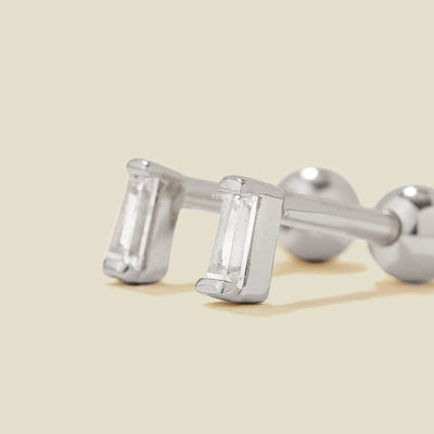 A pair of silver stud earrings with baguette-cut clear gemstones, shown on a solid light beige background.
