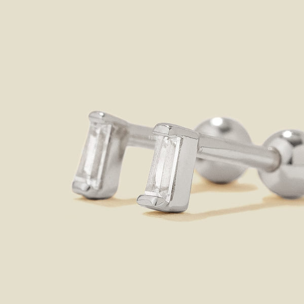 A pair of silver stud earrings with baguette-cut clear gemstones, shown on a solid light beige background.