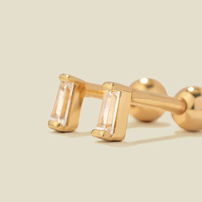 A pair of gold stud earrings with baguette-cut clear gemstones, shown on a solid light beige background.