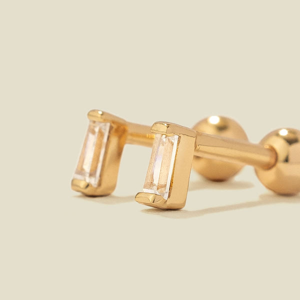 A pair of gold stud earrings with baguette-cut clear gemstones, shown on a solid light beige background.