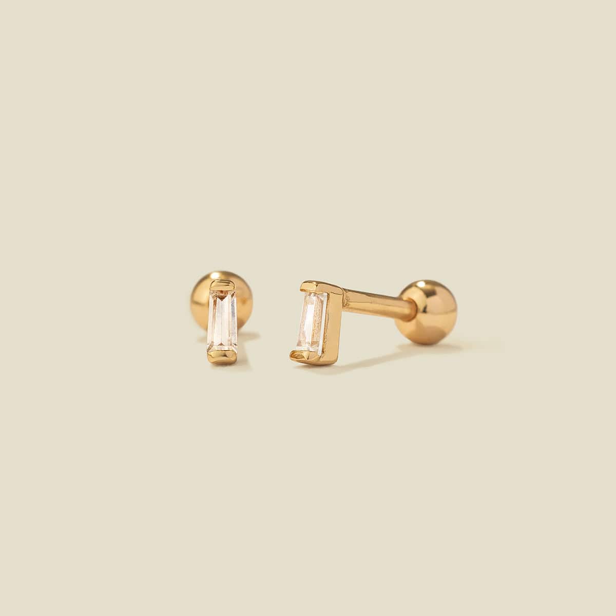 A pair of gold stud earrings with baguette-cut clear gemstones, shown on a solid light beige background.