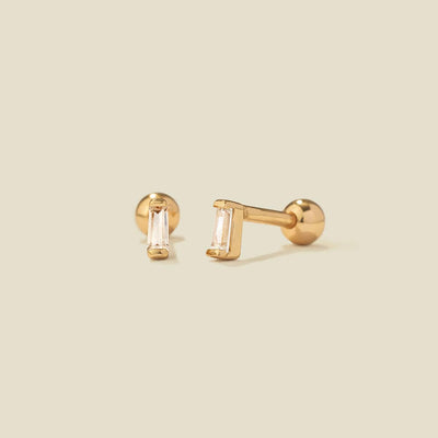 A pair of gold stud earrings with baguette-cut clear gemstones, shown on a solid light beige background.