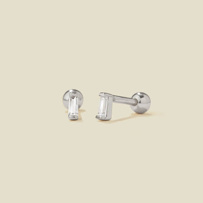A pair of silver stud earrings with baguette-cut clear gemstones, shown on a solid light beige background.