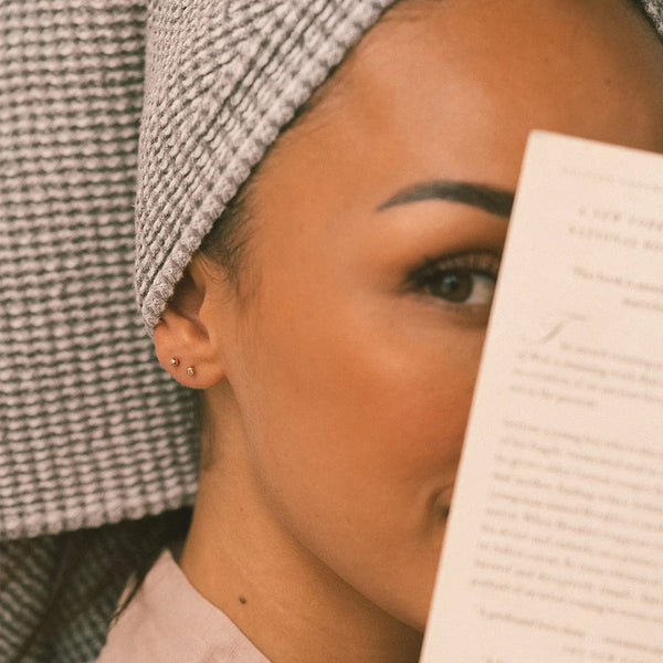 A close-up of a woman with a towel wrapped around her head holding a paper sheet in front of part of her face, wearing two small gold stud earrings.