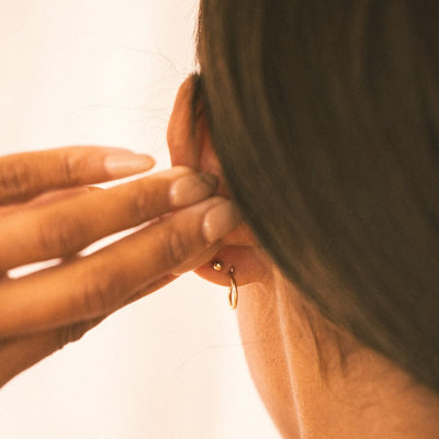 The back of a person's ear wearing a gold thin hoop and a gold round ball backing for a stud.
