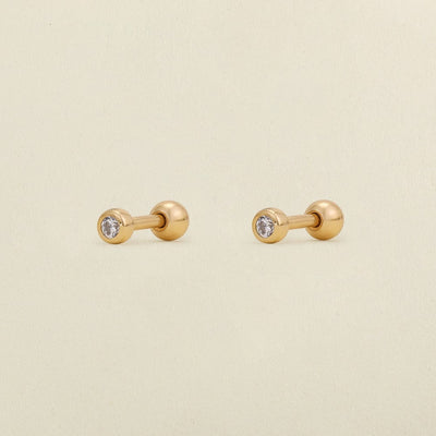 A pair of gold stud earrings with round bezel-set clear gemstones, shown on a light cream textured background.