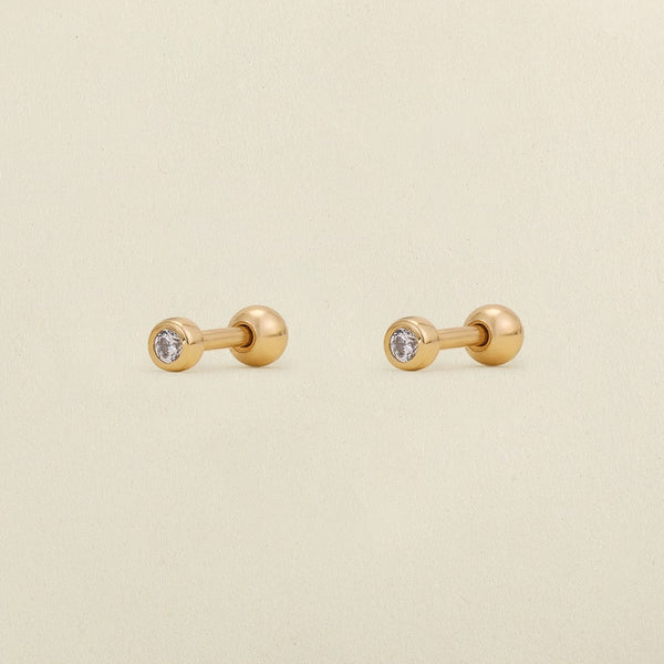A pair of gold stud earrings with round bezel-set clear gemstones, shown on a light cream textured background.