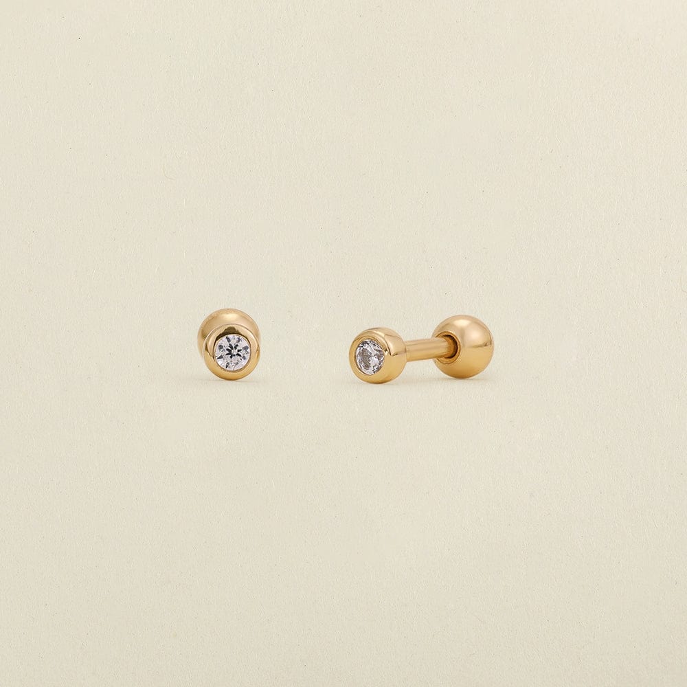 A pair of gold stud earrings with round bezel-set clear gemstones, shown on a light cream textured background.