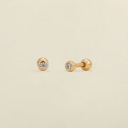 A pair of gold stud earrings with round bezel-set clear gemstones, shown on a light cream textured background.