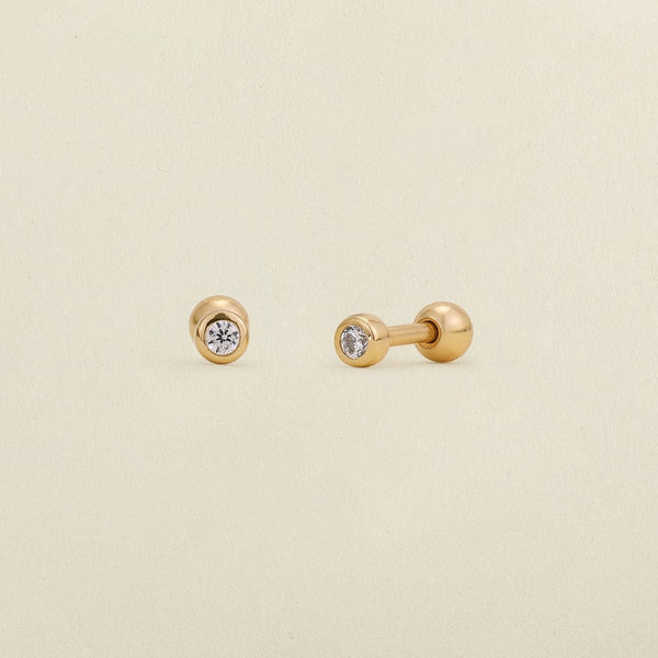 A pair of gold stud earrings with round bezel-set clear gemstones, shown on a light cream textured background.