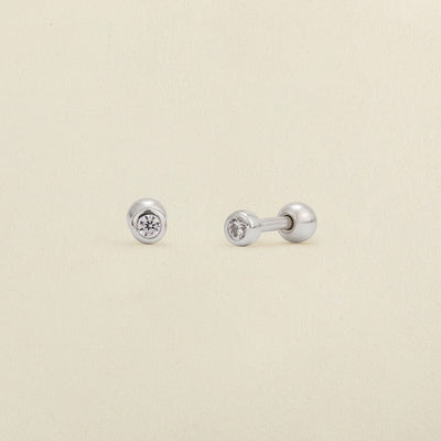A pair of silver stud earrings with round bezel-set clear gemstones, shown on a light cream textured background.