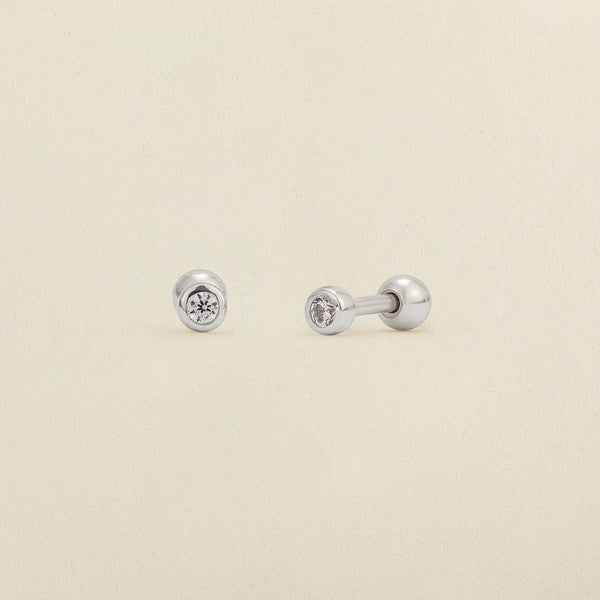 A pair of silver stud earrings with round bezel-set clear gemstones, shown on a light cream textured background.