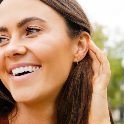 A close-up of a woman's face showcasing her ear wearing a thin gold hoop earring, a gold stud with three bezel-set clear gemstones in a triangle formation, and a single clear bezel-set gemstone stud.