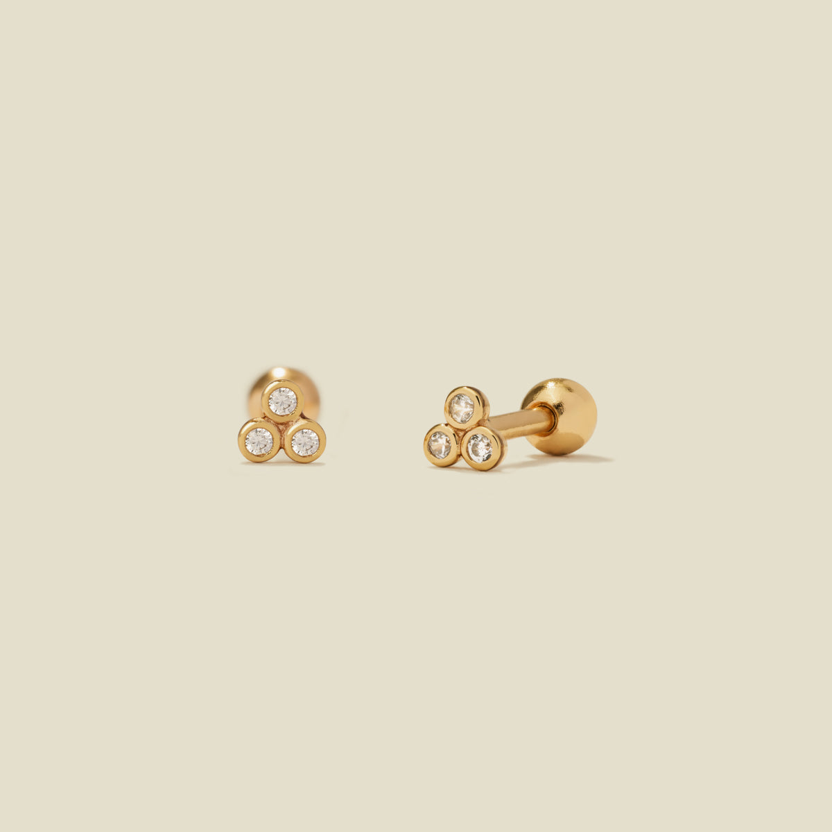 A pair of gold stud earrings with three clear bezel-set gemstones clustered in a triangle on each end, shown on a solid light beige background.