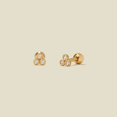 A pair of gold stud earrings with three clear bezel-set gemstones clustered in a triangle on each end, shown on a solid light beige background.