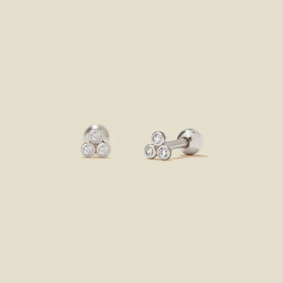 A pair of silver stud earrings with three clear bezel-set gemstones clustered in a triangle on each end, shown on a solid light beige background.