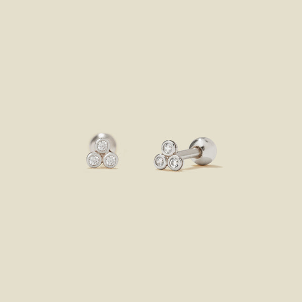 A pair of silver stud earrings with three clear bezel-set gemstones clustered in a triangle on each end, shown on a solid light beige background.