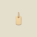 A minimalist gold dog tag pendant with a small row of clear pavé-set stones along the top bail, centered on a solid light beige background.