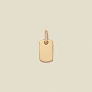 A minimalist gold dog tag pendant with a small row of clear pavé-set stones along the top bail, centered on a solid light beige background.