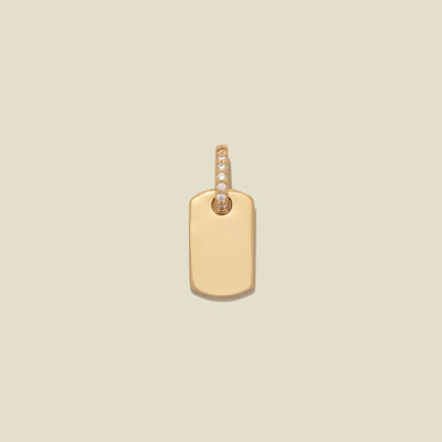 A minimalist gold dog tag pendant with a small row of clear pavé-set stones along the top bail, centered on a solid light beige background.
